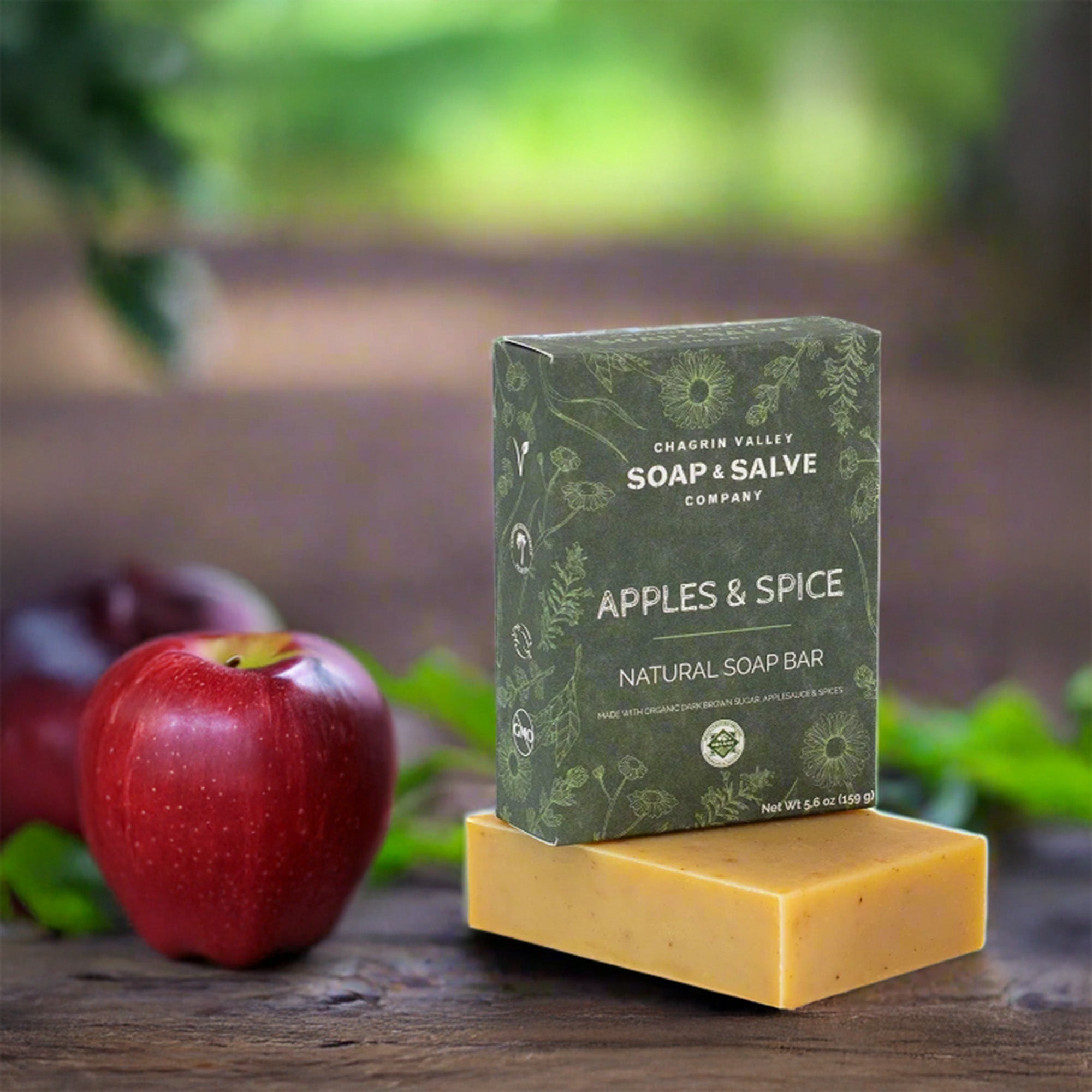Soap bar with 'Apples & Spice' packaging on a wooden surface with apples and greenery in the background.