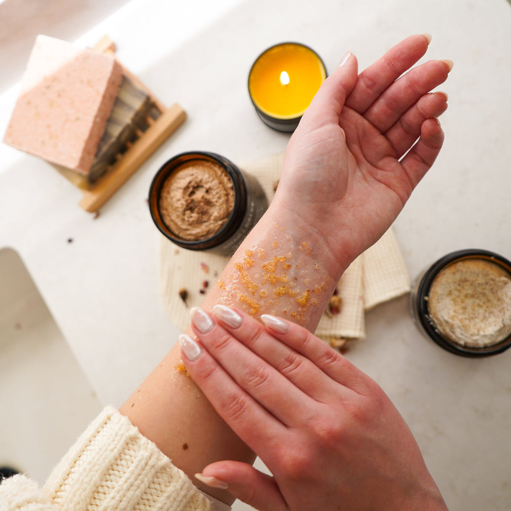 Person applying a scrub to their arm with a candle and soap in the background