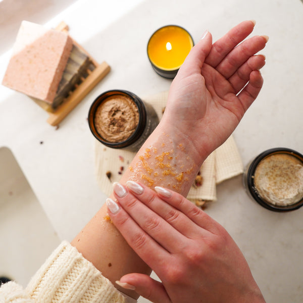 Person applying a scrub to their arm with a candle and soap in the background