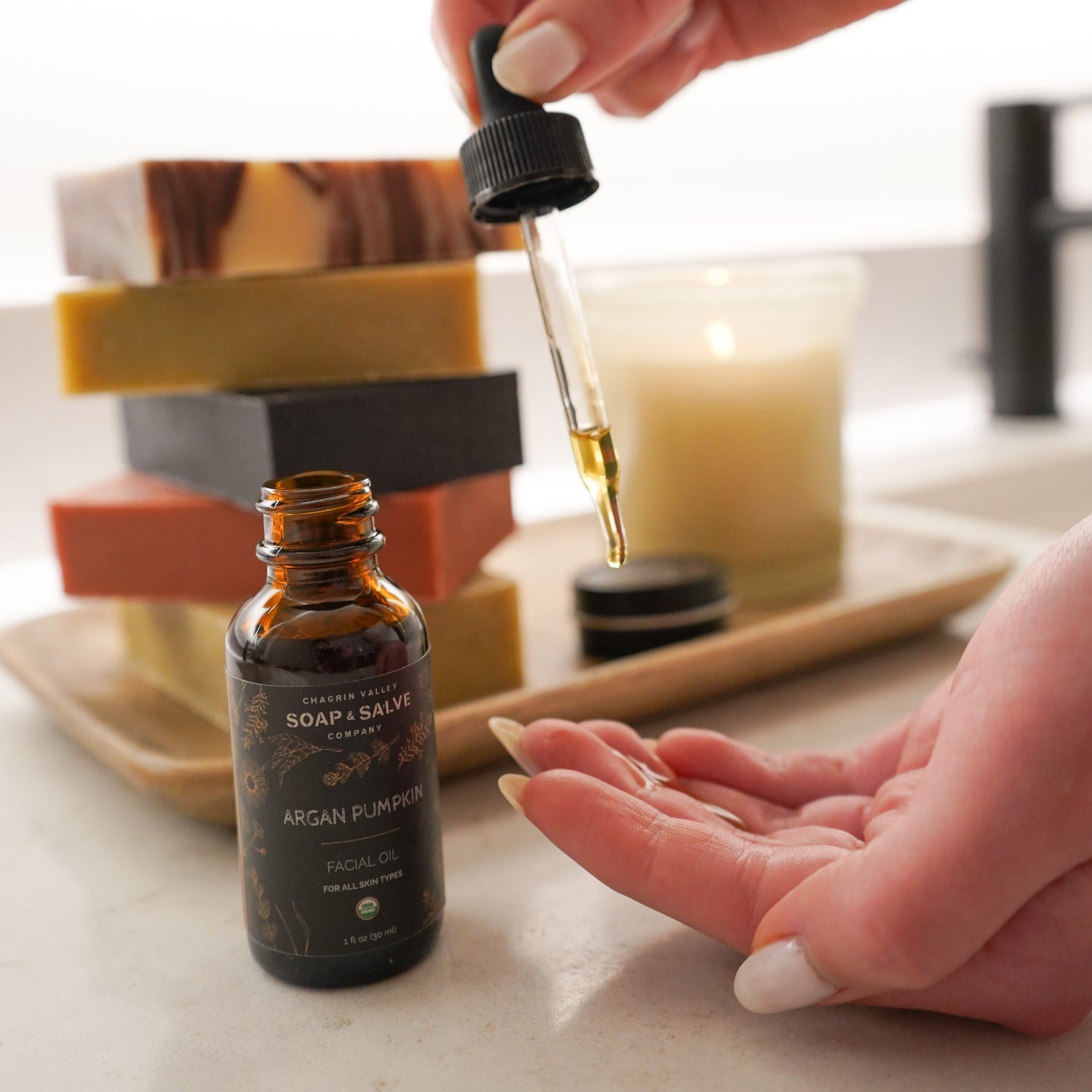 Person using a dropper to apply oil from a bottle on a marble surface with soap and candle in the background.