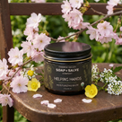 Jar of Chagrin Valley Soap & Salve Company Helping Hands Moisturizing Balm on a wooden surface with flowers.