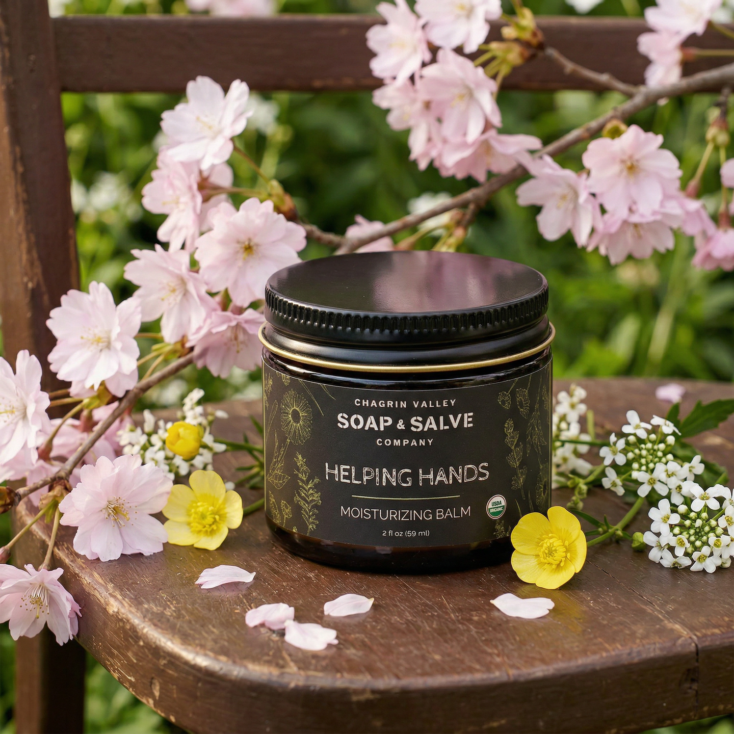 Jar of Chagrin Valley Soap & Salve Company Helping Hands Moisturizing Balm on a wooden surface with flowers.
