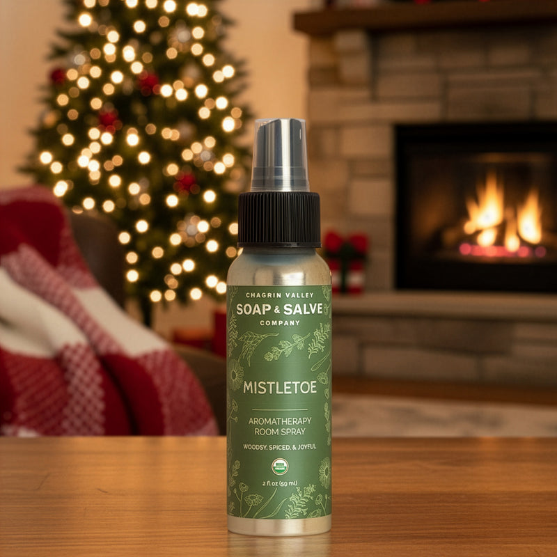 Bottle of Soap & Salve Mistletoe Room Spray on a wooden surface with a Christmas tree and fireplace in the background.