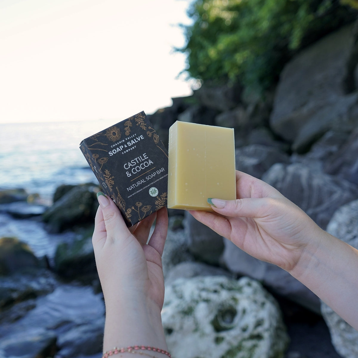 Hands holding the Castile & Cocoa natural soap box and unwrapped bar outdoors by a rocky lakeshore