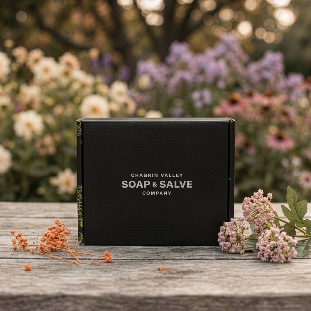 chagrin valley soap and salve monthly subscription box featuring all organic skincare sits on a weathered wooden surface outdoors, with blurred flowers in the background and small dried flowers placed near the box.